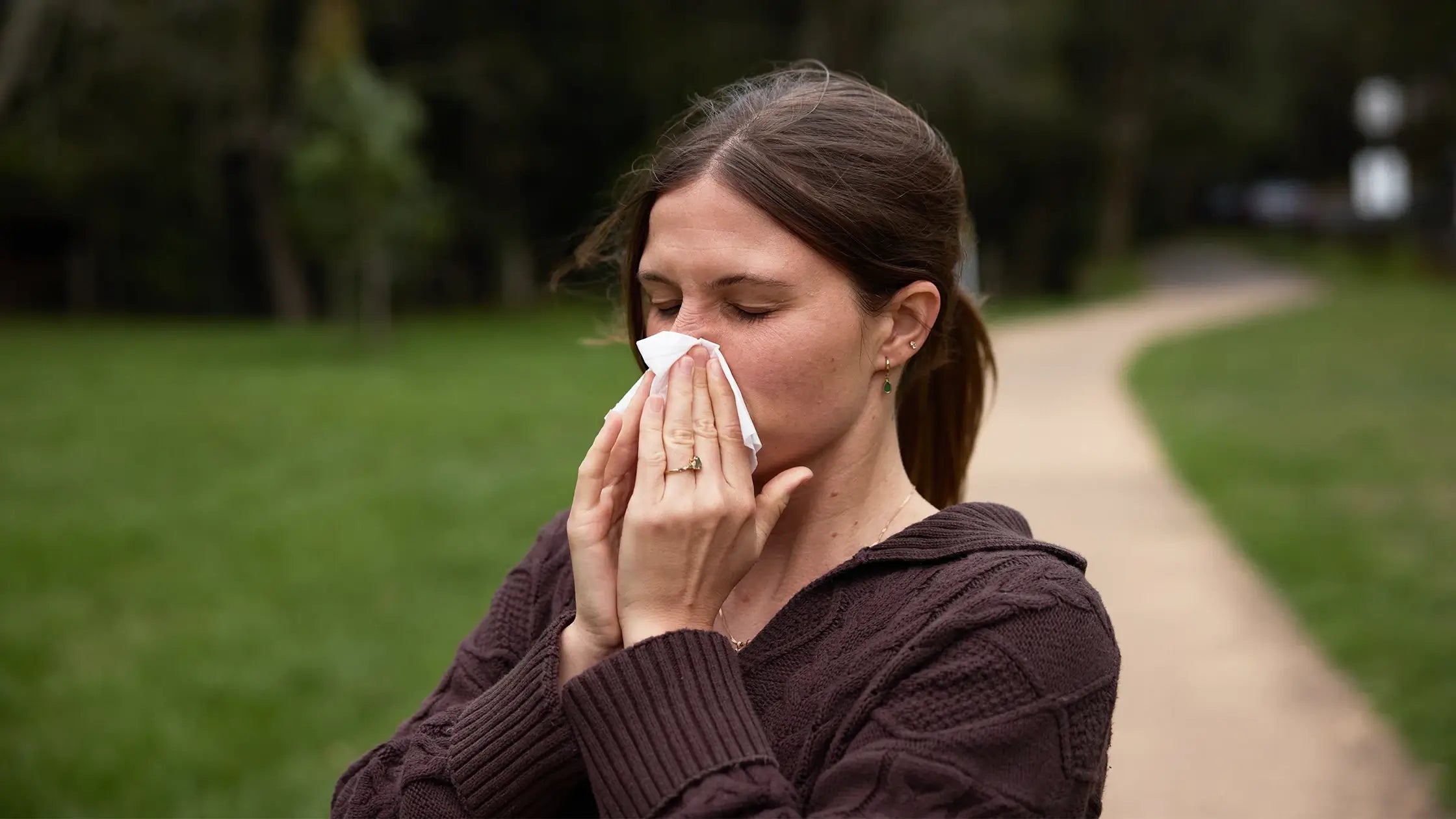 Woman blowing her nose outdoors on a path with greenery.