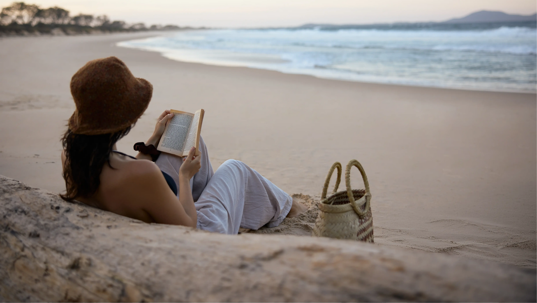 Person reading a book on a sandy beach with a woven bag nearby