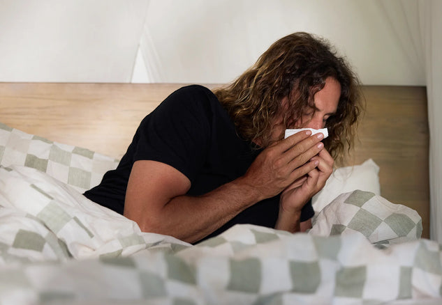 Person lying in bed with a blanket, holding a tissue to their face.
